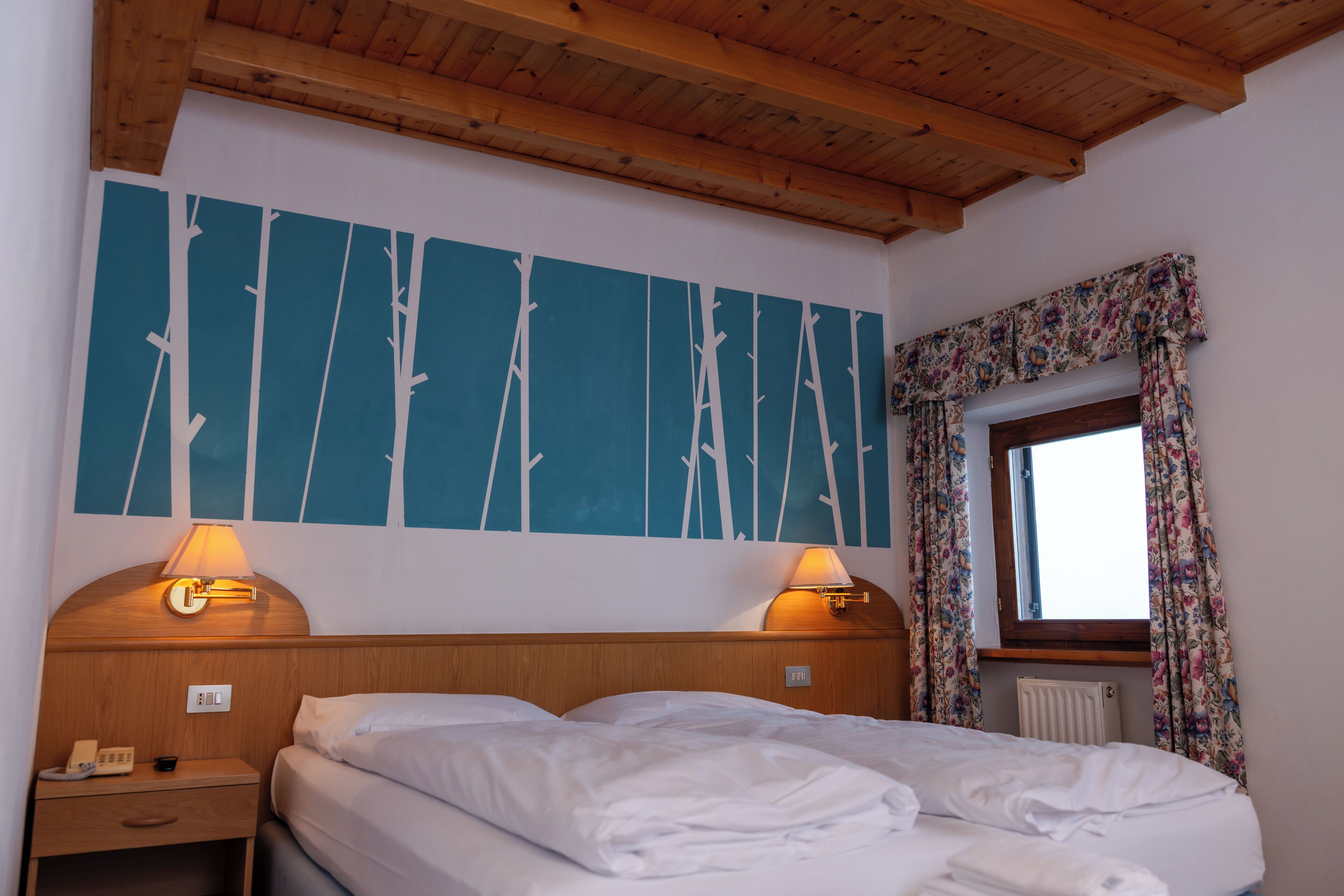 A hotel room with a wooden ceiling, a bed with white linens, two bedside lamps, a wall with a blue abstract design, a window with floral curtains, and a radiator.