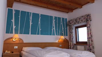 A hotel room with a wooden ceiling, a bed with white linens, two bedside lamps, a wall with a blue abstract design, a window with floral curtains, and a radiator.