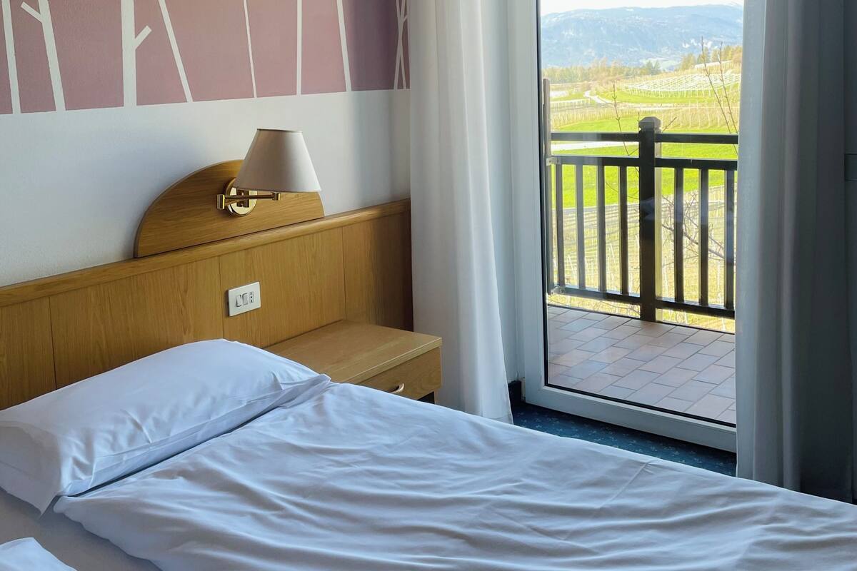 A hotel room with a bed, a wooden nightstand, a lamp, and a view of a balcony with a railing.