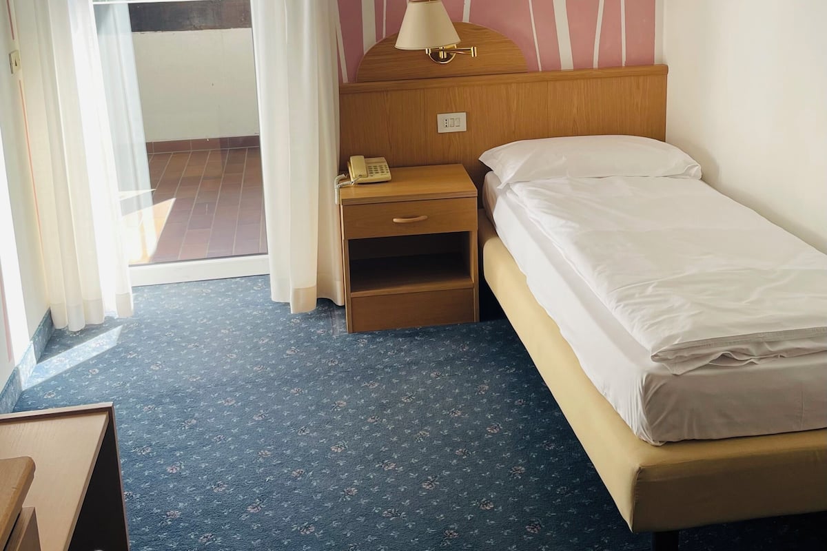 A hotel room with a bed, a bedside table with a lamp, a telephone, and a view of the outdoors.