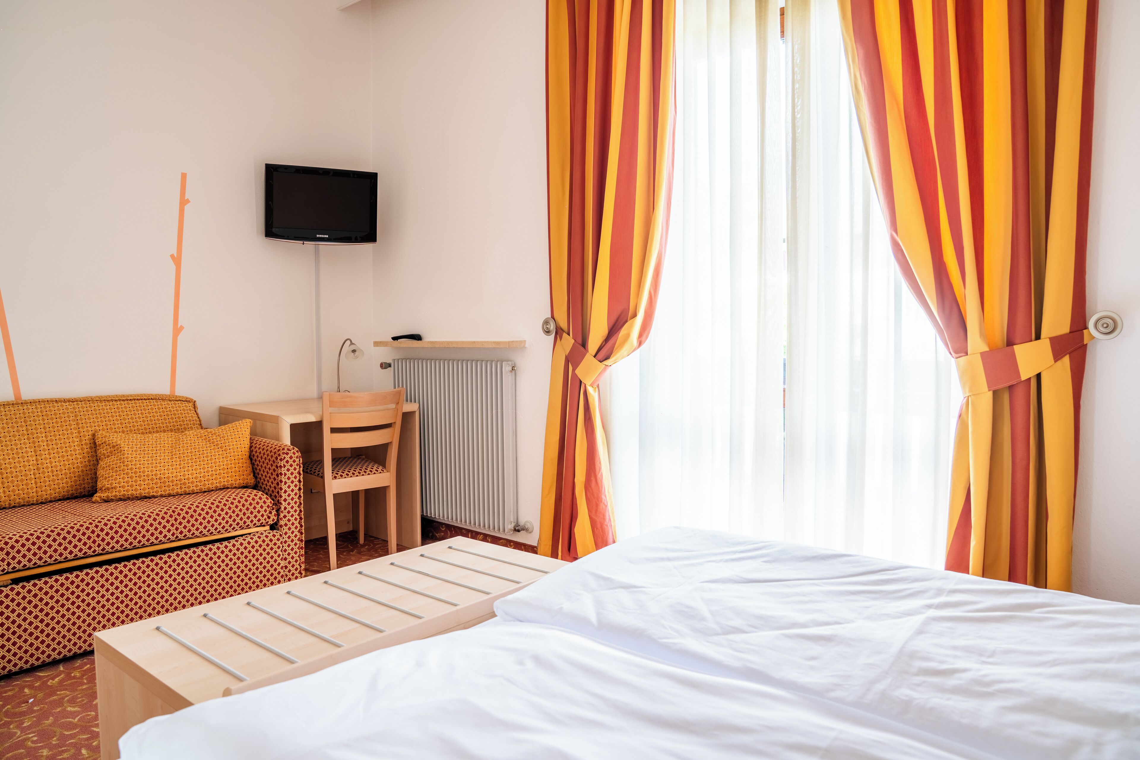A hotel room with a bed, a desk, a chair, a TV mounted on the wall, and a radiator.