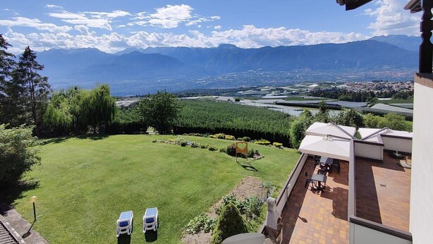A view from a balcony overlooking a lush green lawn, a patio area with outdoor seating, and distant mountains. - Enrosadira (Predaia)
