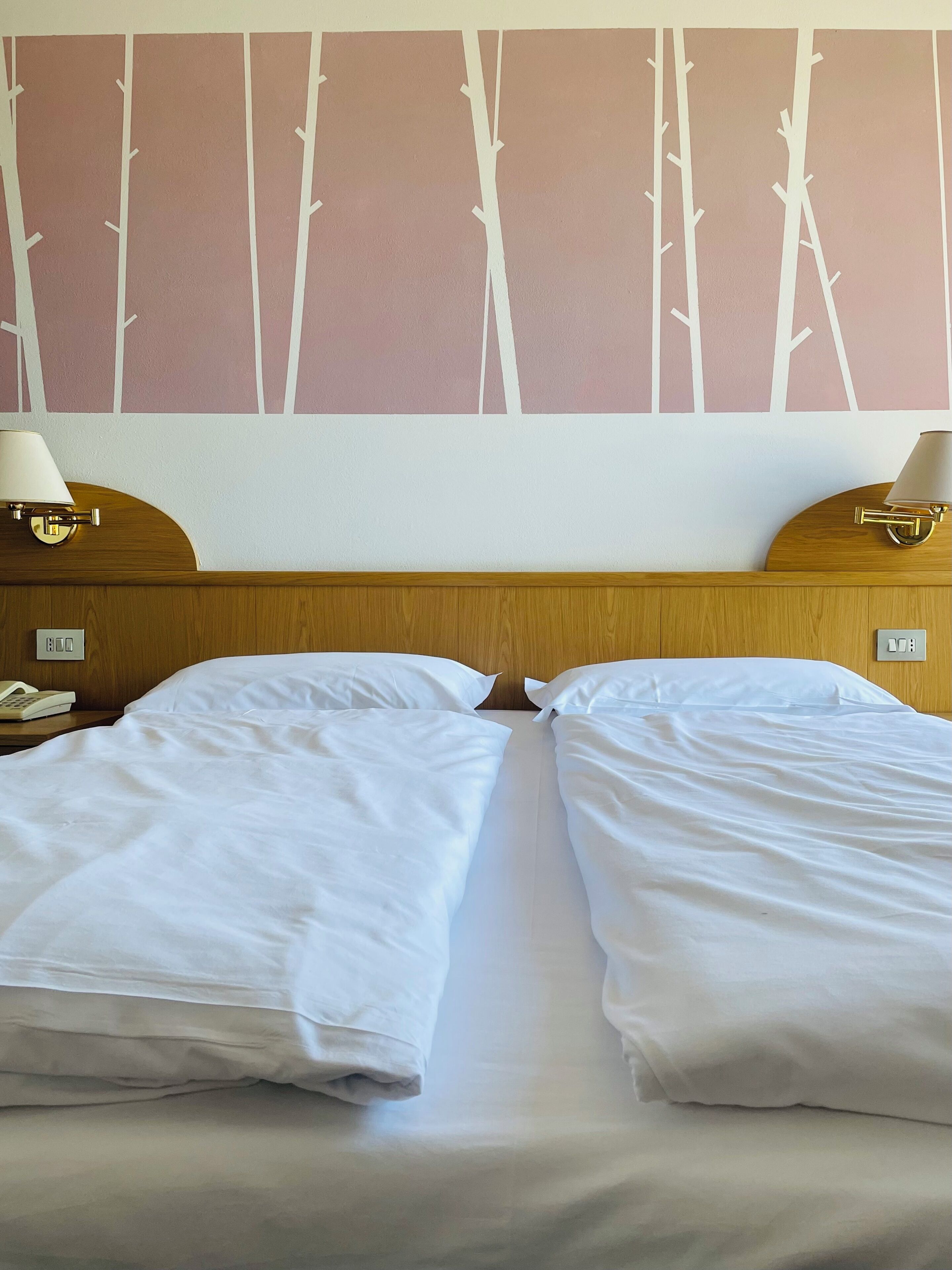 Una habitación de hotel con dos camas individuales, un cabecero de madera, lámparas de noche, un teléfono y una pared decorada con un diseño de árbol.