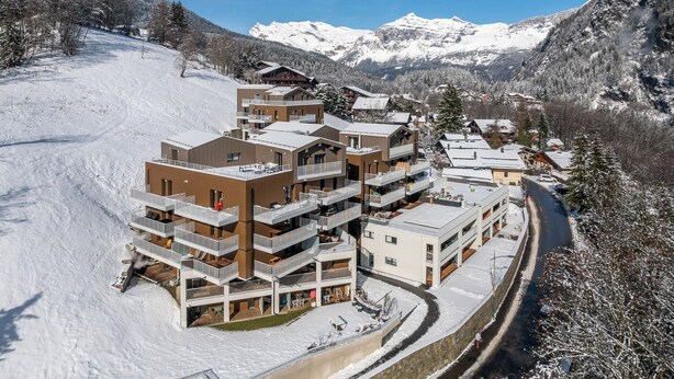 Un bâtiment moderne qui abrite un centre de sports d’hiver, doté de nombreux balcons et entouré de montagnes enneigées sous un ciel dégagé.