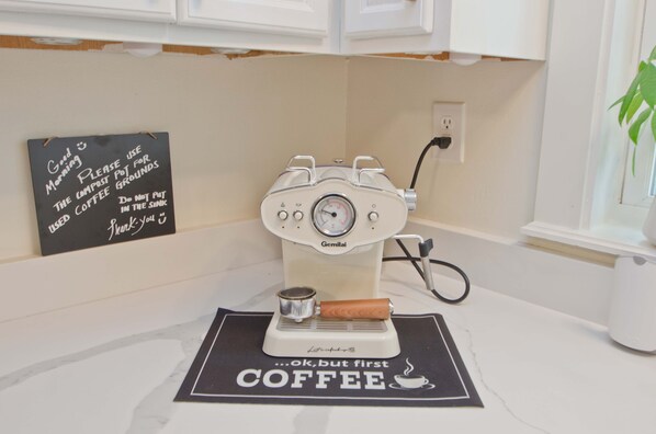 Eine alte Kaffeemaschine steht auf einer Küchenarbeitsplatte, über ihr befindet sich ein Schild und darunter ein Untersetzer mit Kaffeemotiv.