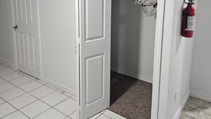 A hallway with white tiled flooring, a closet with hanging clothes, and a fire extinguisher on the wall.