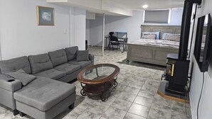 A living room with a grey sofa, a glass coffee table, a bed with patterned bedding, a black heater, and a desk with a chair.