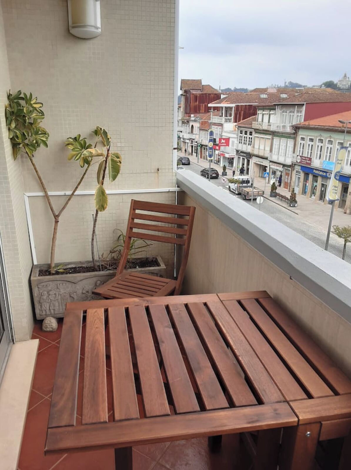 Un balcone con tavolo e sedie in legno, una pianta in vaso e vista su una strada con edifici e negozi.