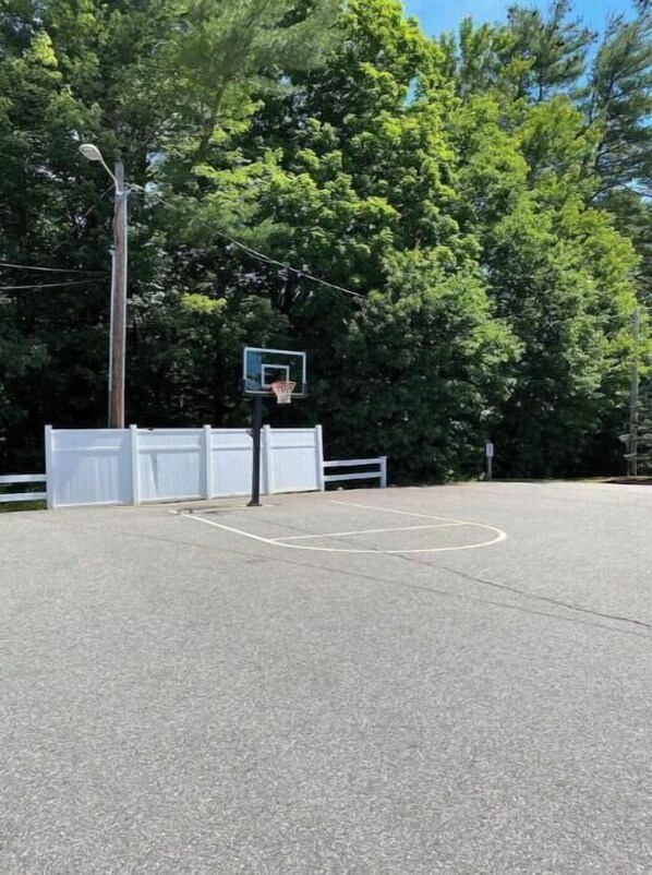Un terrain de basket en plein air, doté d’un panier, entouré d’arbres et délimité par une clôture blanche.