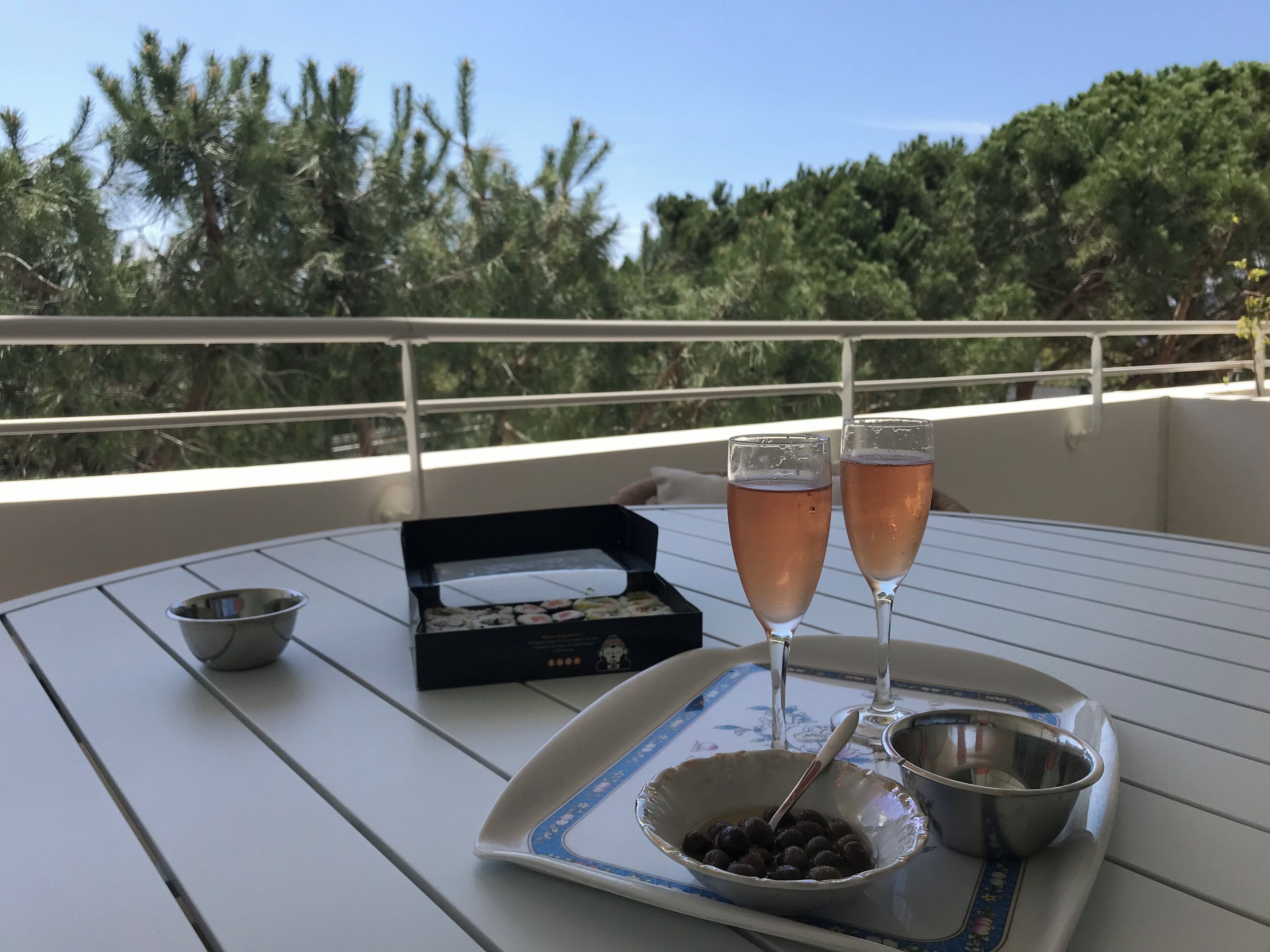 A table with two glasses of a pink beverage, a bowl of chocolate balls, a small bowl, and a box of sushi on a balcony with a railing and trees in the background.
