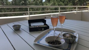 Une table avec deux verres d’une boisson rose, un bol de boules au chocolat, un petit bol et une boîte de sushis, le tout sur un balcon avec une rambarde et des arbres en arrière-plan.