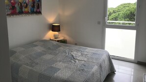 Une chambre à coucher avec un lit, une table de chevet avec une lampe, un tableau au mur et une vue sur la verdure par la fenêtre.