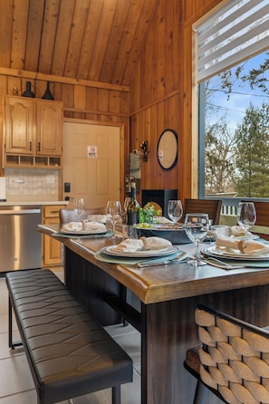 Interior de madera con comedor listo para una comida, con un banco, una mesa con platos y vasos, y vista a árboles a través de una ventana.