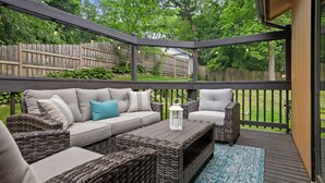 A covered outdoor patio with wicker furniture, a white lantern, and a view of a wooden fence and trees.