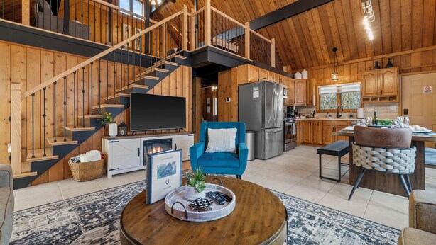A spacious living area with a wooden staircase, a blue armchair, a dining table, and a kitchen in the background.