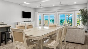 A spacious dining area with a large table and chairs, a television mounted on the wall, and large windows offering a view of the outdoors.