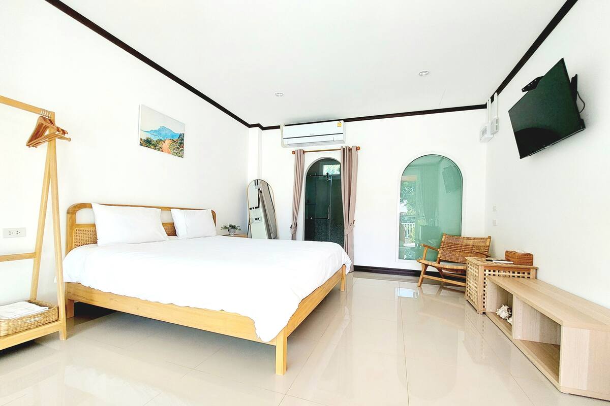 Un dormitorio con cama de madera, televisor montado en la pared, ventana con cortinas y una pequeña mesa con una silla.