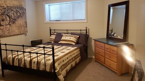 A bedroom with a metal bed frame, a wooden dresser, a large mirror, and a window with blinds. - Walkout Garden 1-bedroom  in Grande Prairie AC, WiFi, laundry, parking space (Grande Prairie)