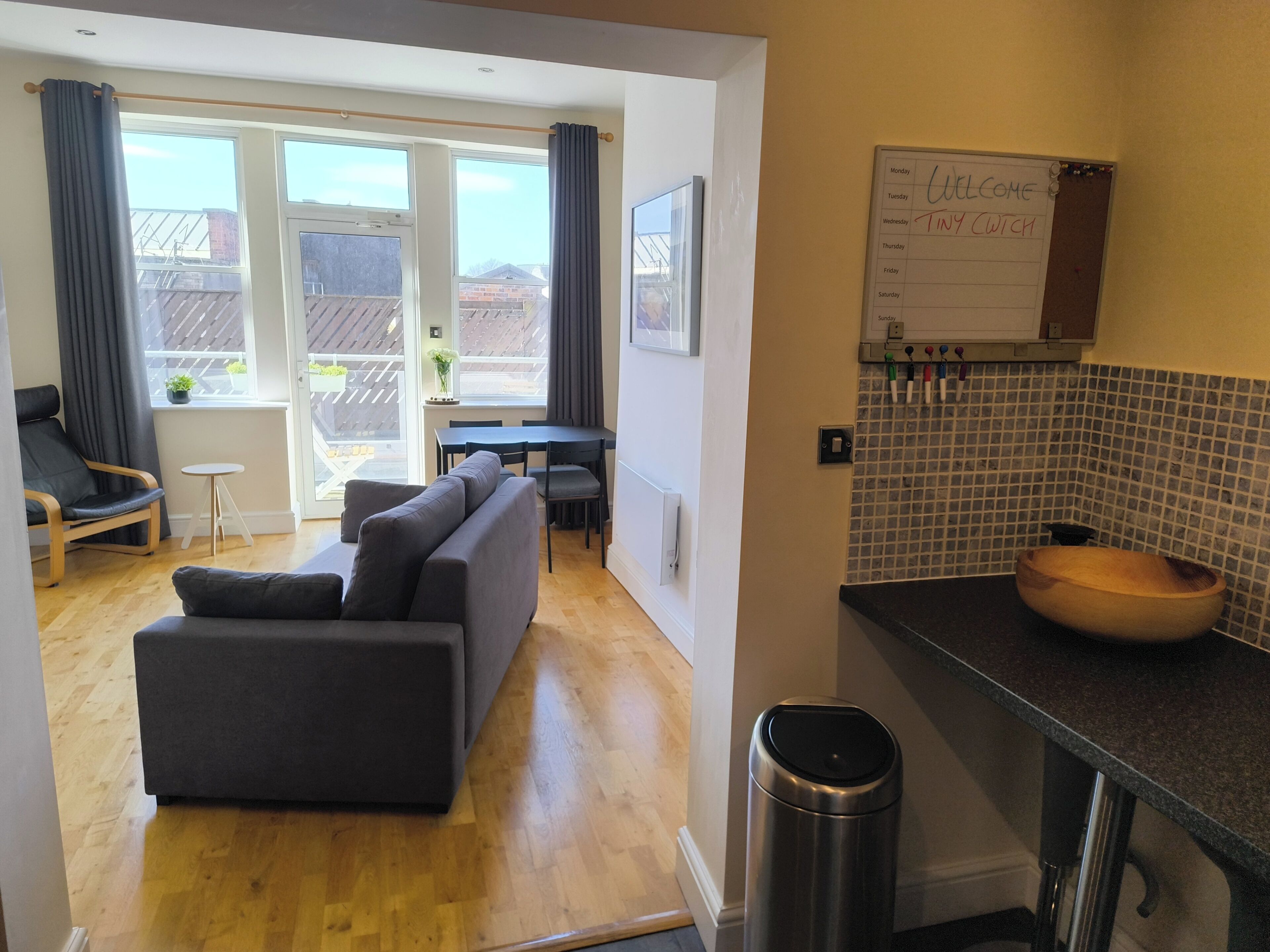 A modern living room with a sofa, dining table, and chairs, a kitchen with a wooden bowl sink, and a whiteboard with a weekly schedule.