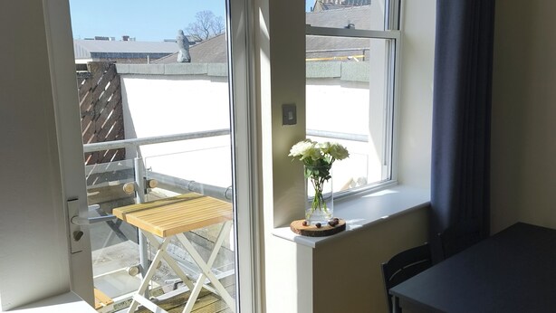 A room with a large window, a wooden table, a chair, and a vase with flowers on a windowsill. - Cardiff Apt - Sleeps 6 - Castle & Stadium Nearby (Cardiff)