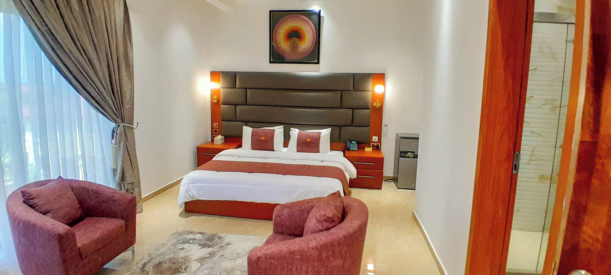 Une chambre à coucher avec un grand lit, deux tables de chevet, une tête de lit, un tableau et deux fauteuils rouges.