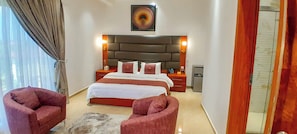 A bedroom with a large bed, two nightstands, a headboard, a painting, and two red armchairs.