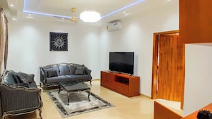 A living room with a sofa, a coffee table, a TV on a wooden stand, and a ceiling light. - Heartland Hotel  (Ada)