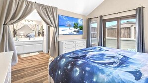 A bedroom with a bed featuring a space-themed bedspread, a large window with a view of a wooden deck, and a painting of a beach scene on the wall.