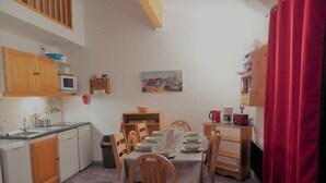 Uma cozinha compacta com armários de madeira, uma mesa de jantar posta para quatro pessoas e uma cortina vermelha à direita.