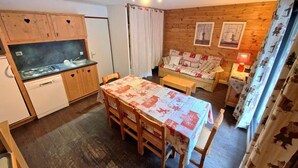 A compact kitchen with wooden cabinets and a built-in dishwasher. There is a dining table with chairs and a small sofa in the living area.