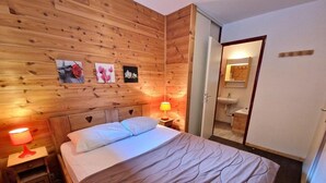 A wooden-paneled bedroom with a bed, two bedside tables with lamps, a mirror, and a small bathroom with a sink and mirror.