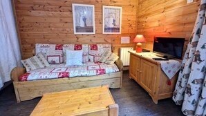 A cozy cabin-style room with a wooden wall, a sofa with patterned cushions, a wooden table, a television on a wooden cabinet, and a fireplace.