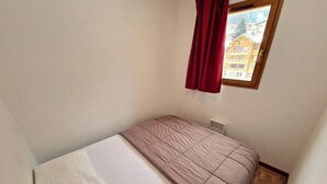 Een kleine kamer met een enkel bed, een houten vloer, een raam met uitzicht op een gebouw en een radiator onder het raam.