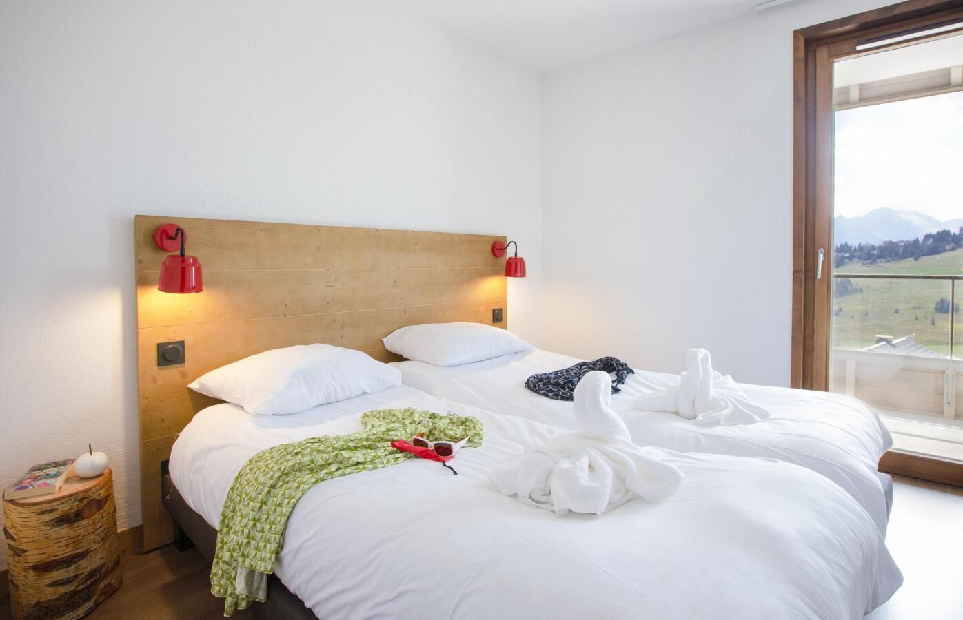 A hotel room with a wooden headboard, two white beds, red wall lamps, a view of mountains through a large window, and a small wooden side table with a candle.