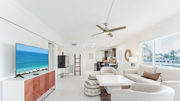 A modern living room with a large flat-screen TV, a ceiling fan, a white sofa, a coffee table, and a view of the ocean through large windows. - Serene Waterfront 2BR 2BA with Intracoastal Views, Pool, Balcony, Parking (Boca Raton)
