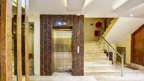 An elevator with a marble wall, a staircase with metal railings, and a fire alarm on the wall. - The York Legacy (New Delhi)