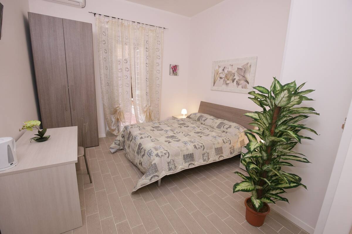 A bedroom with a bed, a bedside table, a potted plant, a curtain, and a wall-mounted artwork.