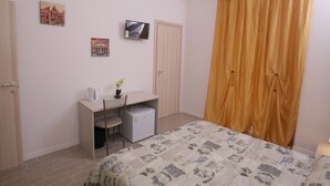 A bedroom with a bed, a desk with a chair, a small refrigerator, a television on the wall, and a large window with curtains. - Rooms Monteverde (Rome)