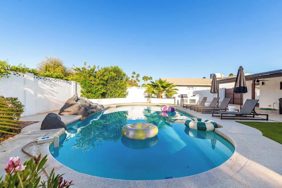 A swimming pool with lounge chairs and umbrellas, surrounded by a white wall and greenery.