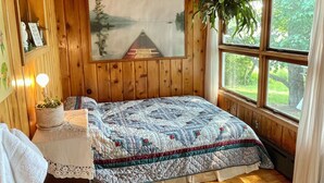 A wooden cabin bedroom with a bed, a window with a view of greenery, a painting on the wall, and a small table with a plant.