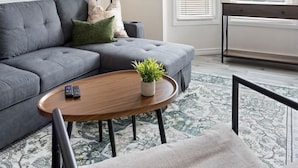 A modern living room with a grey sofa, a round wooden coffee table, a ceiling fan, and a large window with blinds. - Walkerville 1BR Suite Near Caesars Windsor | RiverWalk, Waterfront & Downtown (Windsor)