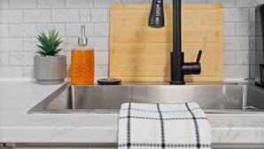 A kitchen with a black faucet, white cabinets, a marble backsplash, a wooden cutting board, a potted plant, and a towel hanging on the sink. - Walkerville 1BR Suite Near Caesars Windsor | RiverWalk, Waterfront & Downtown (Windsor)
