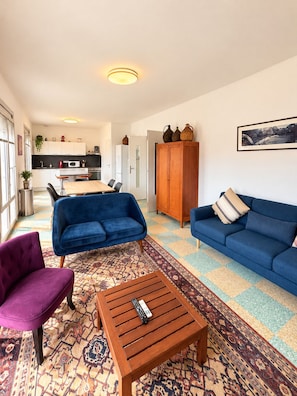 Ein Wohnzimmer mit einer Couch, einem Sessel und einem Couchtisch, im Hintergrund eine Küche und ein großer Holzkasten.