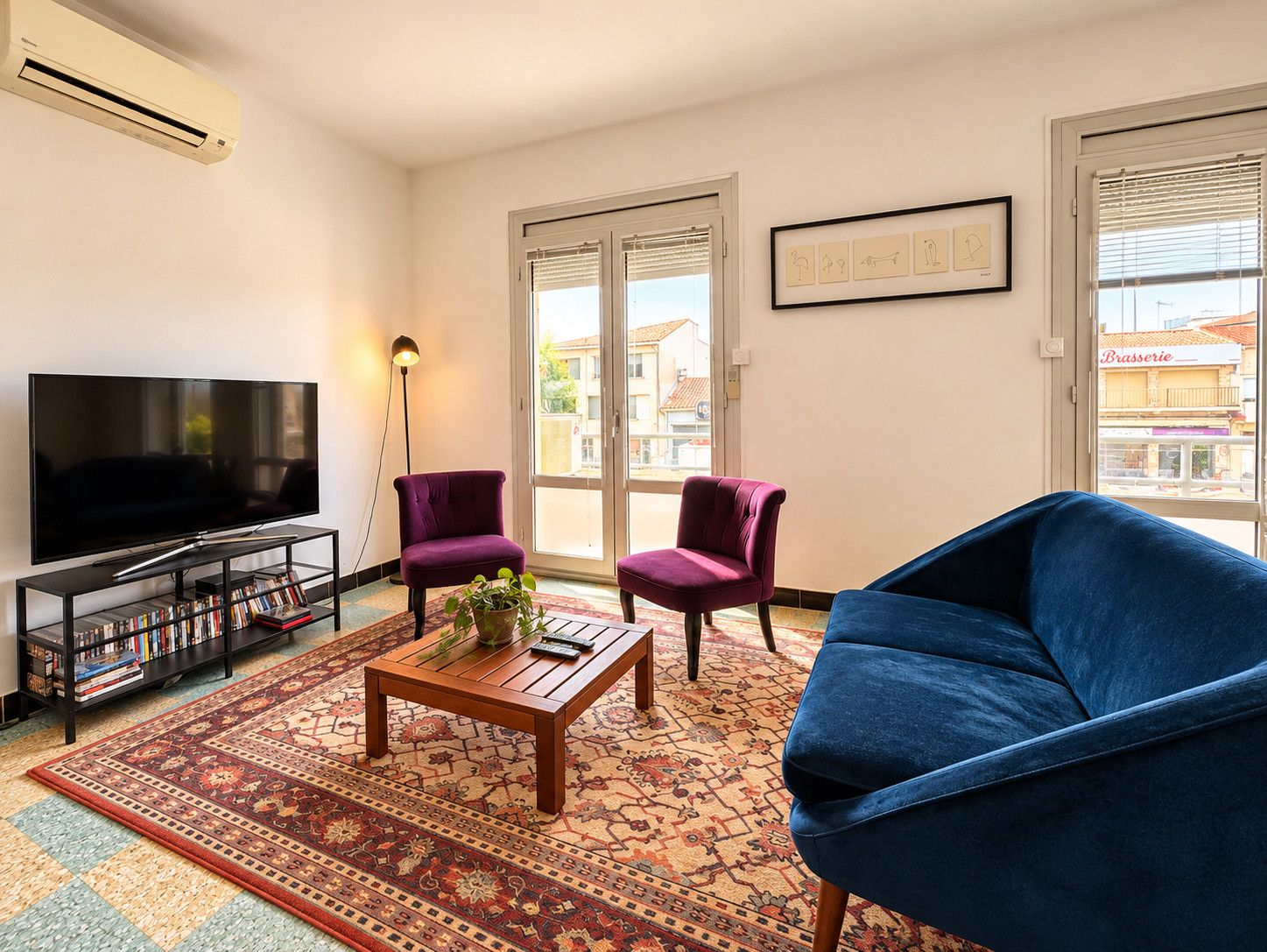 Uma sala de estar com televisão de tela plana, duas poltronas roxas, um sofá azul, uma mesa de centro de madeira e um tapete estampado.
