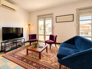 Ein Wohnzimmer mit einem Flachbildfernseher, zwei violetten Sessel, einem blauen Sofa, einem Holztisch und einem gemusterten Teppich.