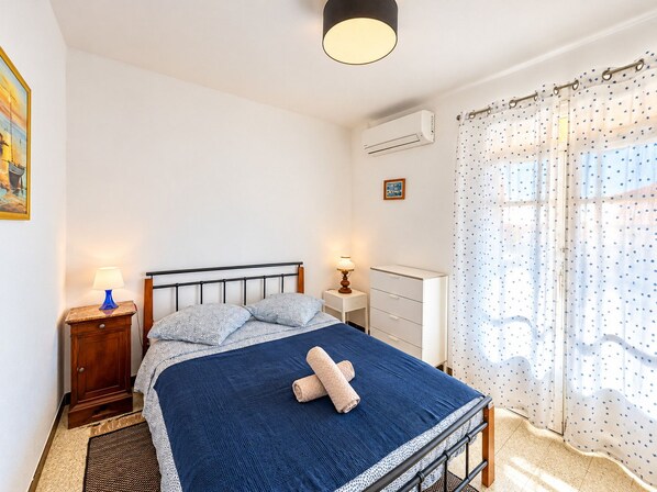 Un dormitorio con cama, mesitas de noche, lámpara, unidad de aire acondicionado, ventana con cortinas y un cuadro en la pared.
