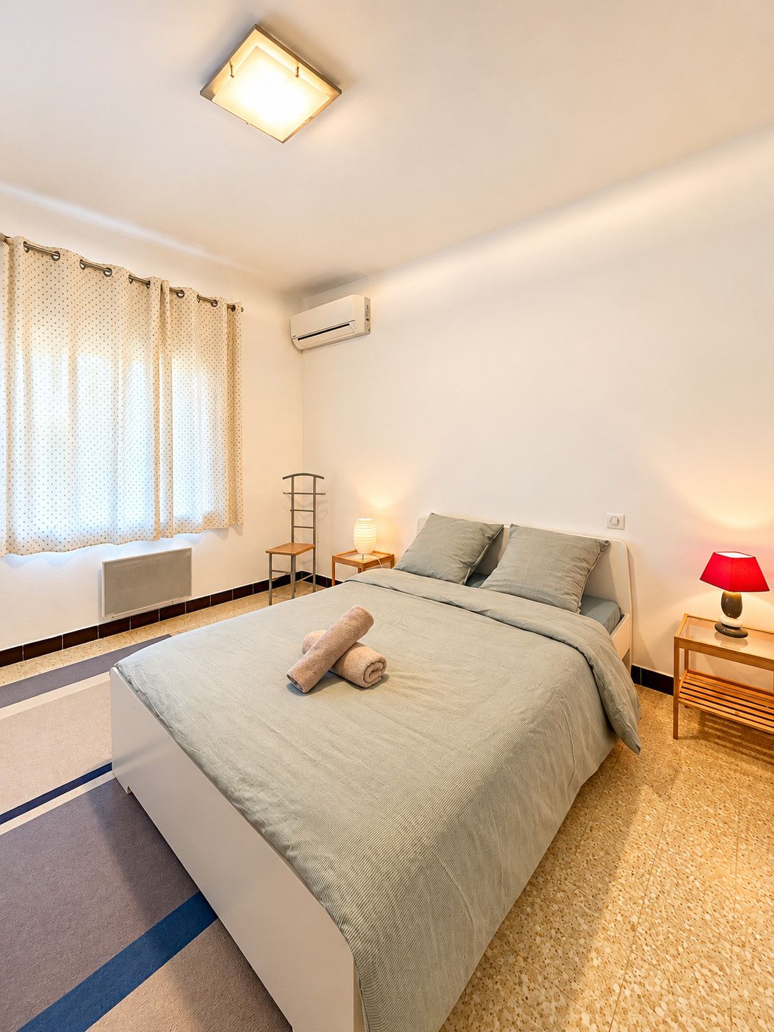 Een hotelkamer met een bed, een nachtkastje met een lamp, een raam met gordijnen, een airconditioning en een radiator.