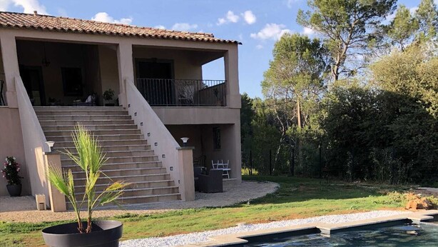 A swimming pool with a stone edge, a potted palm plant, and a two-story house with a balcony. - Cocon d'Amour & Spa (Le Thoronet)