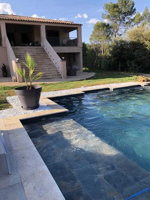 Una piscina con bordo in pietra, una pianta di palma in vaso e una casa a due piani con un balcone.