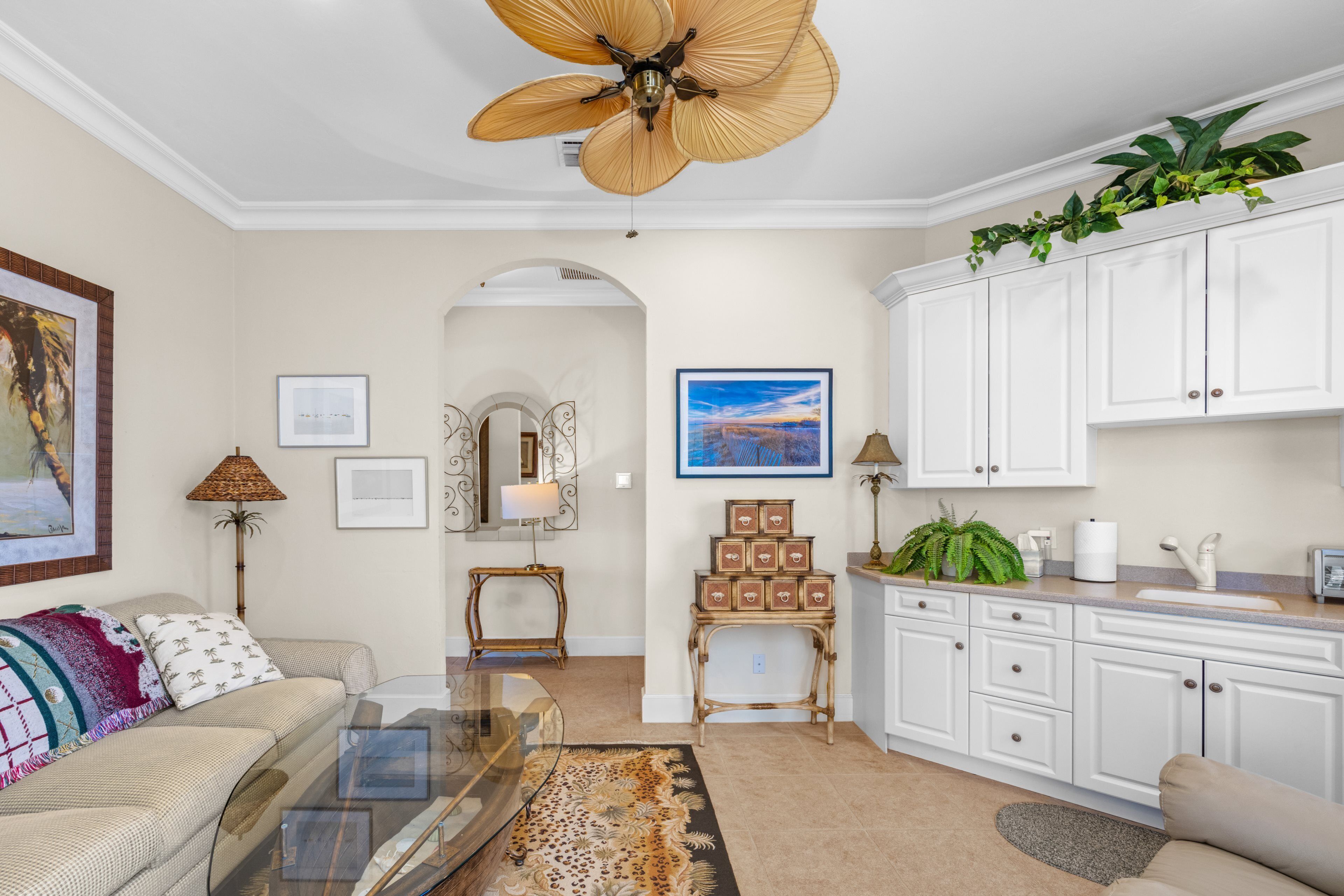 A living room with a glass coffee table, a sofa, a ceiling fan, a dining area with white cabinets, and a kitchen with a sink and countertop.